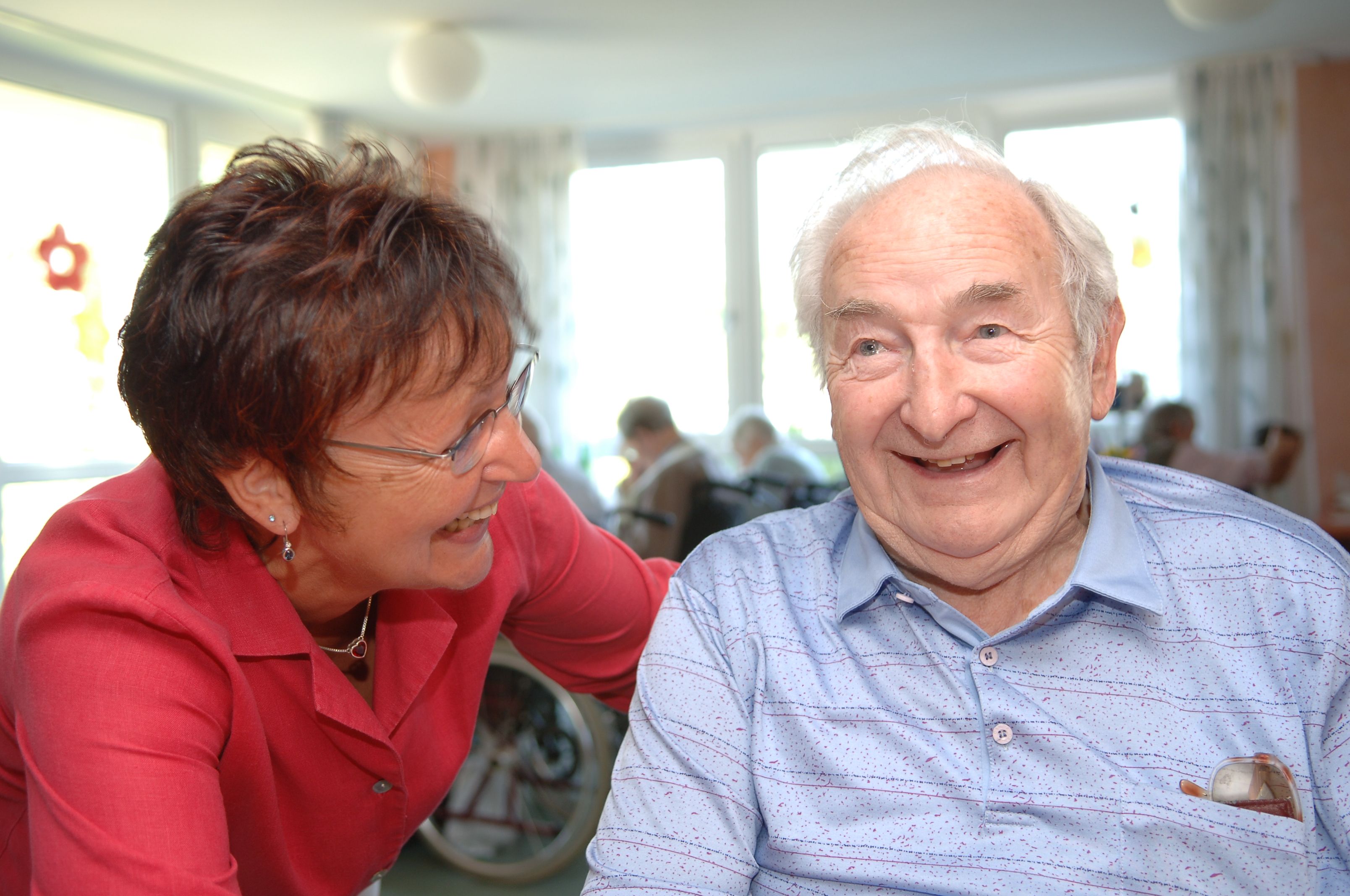 Eine ASB-Mitarbeiterin steckt mit einem Senior die Köpfe zusammen,beide lächeln